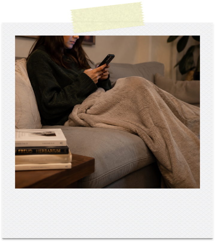 A woman scrolling her phone under a blanket instead of reading the books beside her